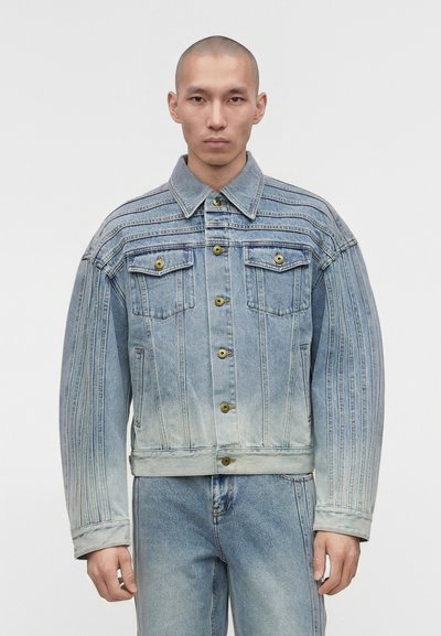 Homme à la tête rasée portant une veste en denim bleu clair avec des coutures verticales et un jean assorti sur fond blanc.