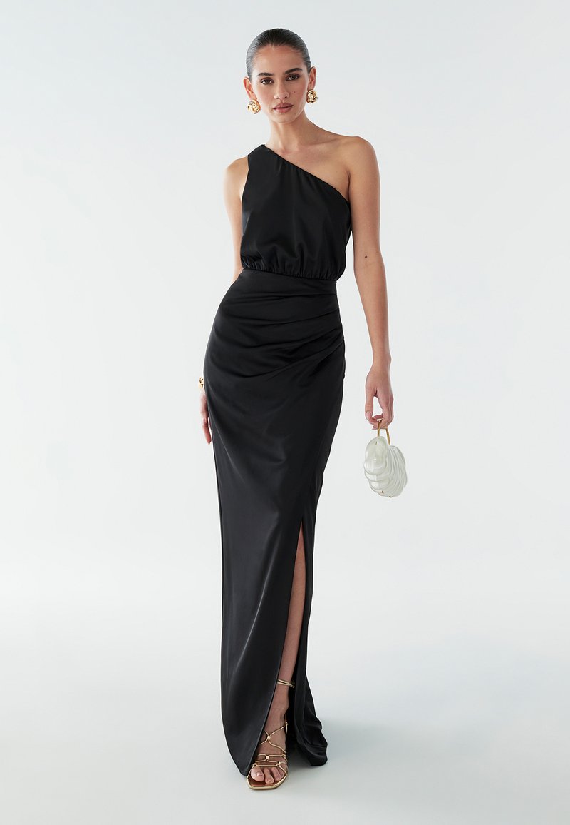 Melnas satīna vakartērps ar vienu plecu, savilktu vidukli un augstu šķēlumu līdz augšstilbam; apvienots ar baltu rokassomu un zeltainām sandālēm ar siksniņām.