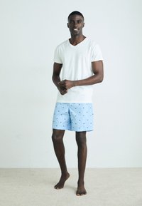 White short-sleeve V-neck shirt paired with light blue swim shorts featuring a pattern of small dark blue logos. Model stands barefoot on a carpet.