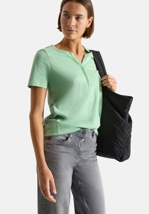 Femme portant un haut vert clair à manches courtes et un jean gris, tenant une veste matelassée noire sur son épaule, regardant légèrement sur le côté.