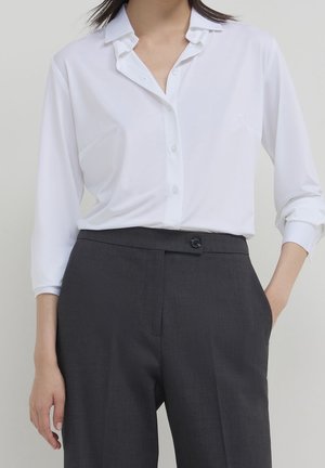 Vrouw draagt een witte blouse met knopen, ingestopt in hooggesneden donkergrijze broek, met één hand in de zak tegen een effen achtergrond.