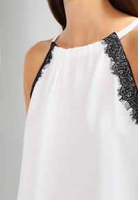 Top halter blanco con acentos de encaje negro en las correas. Tela suave y diseño fluido, con delicado ribete de encaje a lo largo de los hombros.