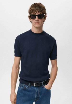 Jersey de manga corta de color navy con cuello redondo, combinado con jeans de color azul claro y un cinturón negro. El modelo lleva gafas de sol negras.
