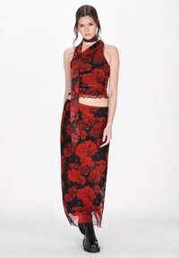 Fekete anyagú ruha piros rózsa mintával. Cropped top nyakkendővel és fodros szegéllyel, hosszú szoknyával párosítva. Fekete lábbeli látható.