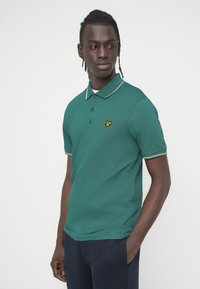 Πράσινο πόλο μπλουζάκι με λευκό και γκρι ριγέ γιακά, δύο κουμπιά στη λαιμόκοψη και κίτρινο έμβλημα πουλιού στο στήθος. Κοντά μανίκια.