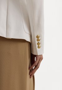 Manche d'un blazer blanc texturé présentant quatre boutons décoratifs dorés, assortie à un matériau lisse couleur sable en dessous.