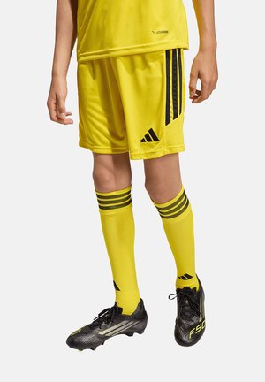 Person trägt gelbe Adidas-Fußballshorts, passende gelbe Socken mit schwarzen Streifen und schwarze Fußballschuhe mit gelben Akzenten.