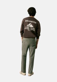 Hnědá mikina s bílým textem a grafikou hor, doplněná světle zelenými kalhotami. Oblečení má ležérní, jednoduché linie a jemné textury.