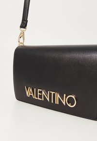 Melns rokas somas no gludas sintētiskās vielas ar zelta "VALENTINO" logo, aprīkota ar noņemamu siksnu un taisnstūrveida formu.
