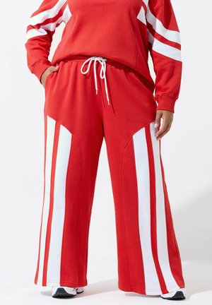 Person iført røde træningsbukser med brede hvide lodrette striber ned langs benene og en snøre i taljen, kombineret med en matchende rød og hvid top.