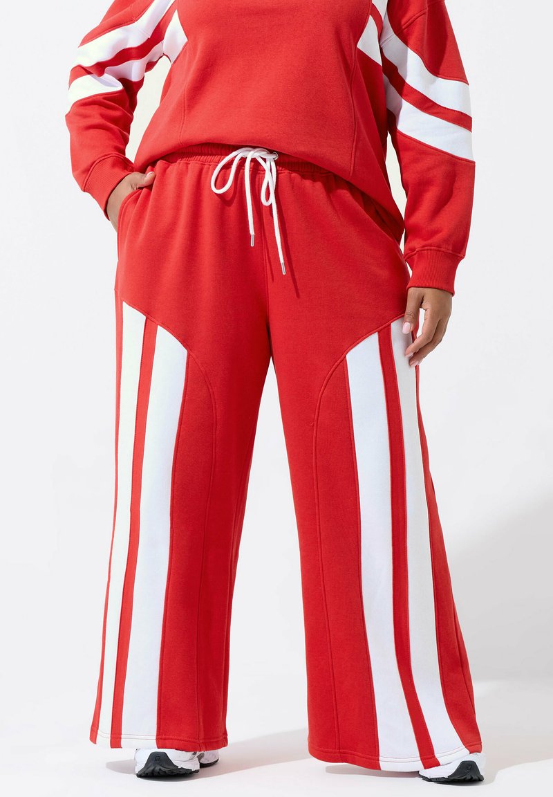 Person trägt rote Jogginghosen mit breiten weißen vertikalen Streifen entlang der Beine und einem Bündchen mit Kordelzug, kombiniert mit einem passenden roten und weißen Oberteil.