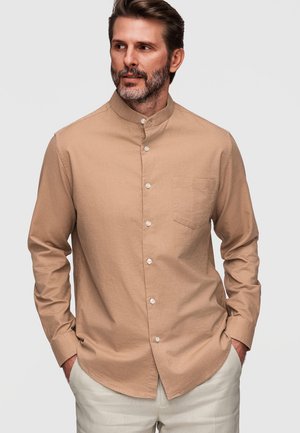 Homme avec barbe portant une chemise beige à col mandarin boutonnée et un pantalon clair, les mains dans les poches, regardant sur le côté.