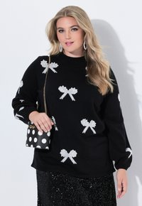 Czarny sweter z białymi wzorami w kokardki, okrągły dekolt i długie rękawy. Modelka trzyma torbę w czarno-białe kropki. Widoczna spódnica z cekinami.