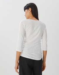 Witte, getailleerde top met 3/4 mouwen en een ronde halslijn. Heeft een middennaad op de rug en een zachte, gladde textuur.