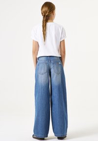 Jeans de denim azul de pierna ancha con un acabado desgastado, combinados con una camisa blanca de manga corta. La tela parece suave, con detalles de costura visibles.