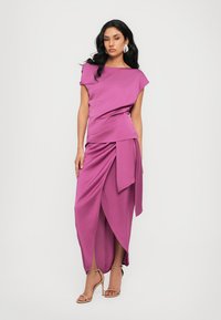 Femme portant une robe enveloppante sans manches magenta avec un drapé asymétrique et des sandales à talons nus sur un fond blanc.