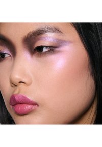 Primer plano de un rostro con sombra de ojos púrpura iridiscente, labios brillantes en rosa y piel suave con un sutil brillo en la mejilla.