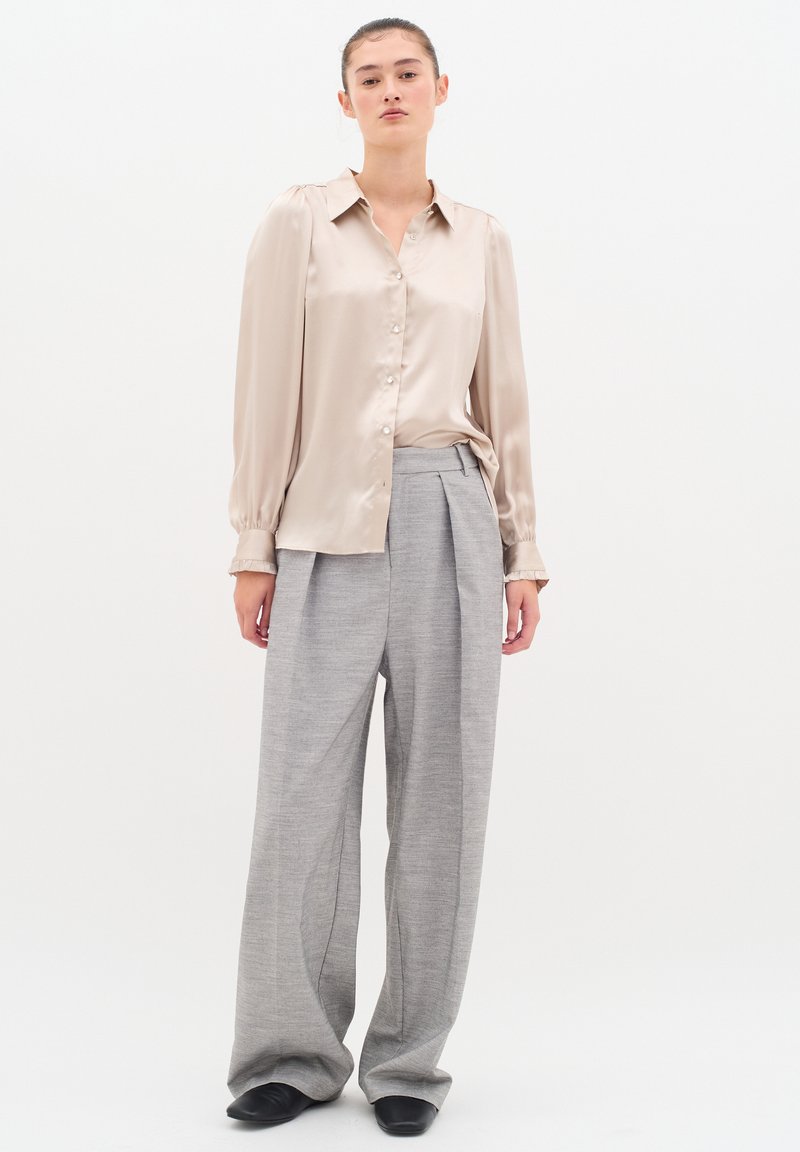 Blusa satin beige con chiusura a bottoni e maniche a sbuffo; abbinata a pantaloni grigi a gamba larga con pince e tessuto testurizzato. Scarpe nere.