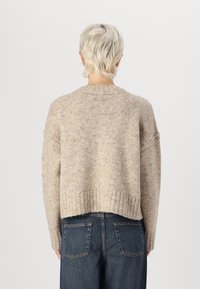 Osoba z krótkimi jasnymi włosami, ubrana w beżowy sweter dzianinowy i niebieskie dżinsy, stojąca tyłem na jednolitym jasnym tle.