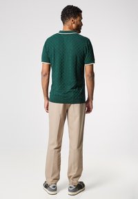 Camiseta polo verde texturizada con mangas cortas, detalles en blanco, combinada con pantalones beiges y zapatillas grises. Vista trasera del atuendo.