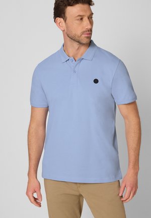 Mężczyzna ubrany w jasnoniebieską koszulkę polo z krótkim rękawem i dwoma guzikami oraz beżowe spodnie, patrzący w lewo na jednolitym tle.