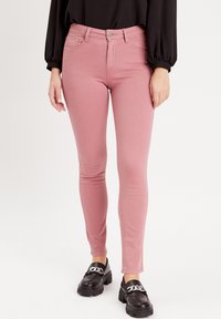 Jeans skinny stretch rose avec cinq poches et une fermeture à bouton à l'avant, associés à des mocassins noirs avec un accent de chaîne décoratif.