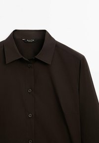 Chemise marron foncé à manches longues avec boutons et col classique, présentée sur un fond blanc uni.