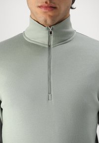 Suéter de cremallera verde claro con un cuello alto, textura suave y una cremallera corta, enfatizando un diseño minimalista.