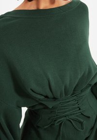 Dunkelgrüner, gerippter Pullover mit weitem Ausschnitt und gerafften Details am Rücken. Verfügt über Schnürungen für eine verstellbare Passform. Weiche Textur.