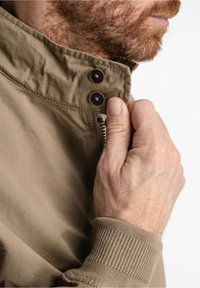Khaki jacket with a high collar, featuring copper buttons and ribbed cuffs. Material appears smooth with a subtle sheen. Zippered front detail.
