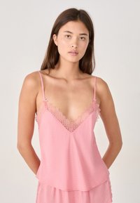 Modelo con cabello largo castaño lleva una blusa sin mangas rosa con encaje. Ella se encuentra de pie contra un fondo neutro, con las manos relajadas a los lados.