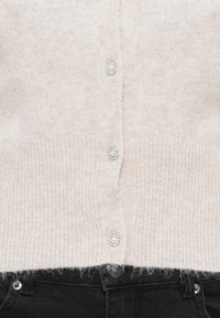 Cárdigan de punto color beige claro con una textura suave, cinco botones adornados con cuentas y un dobladillo acanalado, combinado con jeans negros.