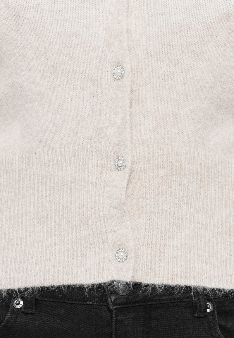 Helles beige Strickcardigan mit weicher Textur, fünf mit Perlen besetzten Knöpfen und geripptem Saum, kombiniert mit schwarzen Jeans.