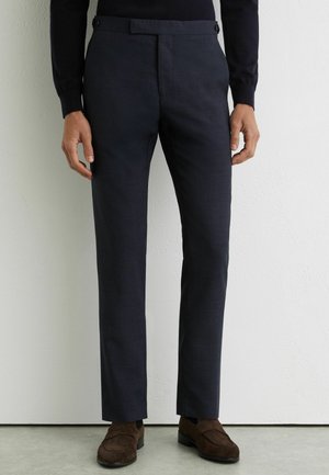 Mężczyzna w granatowych, dopasowanych garniturowych spodniach z bocznymi regulatorami i brązowych zamszowych mokasynach, stojący przy białej ścianie.