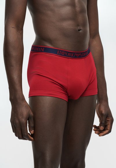 Férfi törzs piros Emporio Armani boxerrövidnadrágot visel, sötétkék derékpánttal, tiszta világos háttér előtt áll.