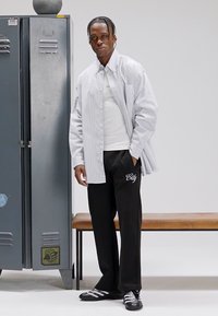 Ριγέ πουκάμισο με κουμπιά, λευκό T-shirt, μαύρο παντελόνι με κέντημα "City" και μαύρα αθλητικά παπούτσια. Στο φόντο εμφανίζεται ένα μεταλλικό ντουλάπι και ένα ξύλινο παγκάκι.