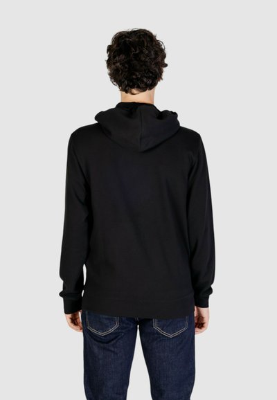Felpa nera con cappuccio realizzata in un morbido tessuto testurizzato. Presenta un design semplice con orlo e polsini a costine, abbinata a jeans di denim.