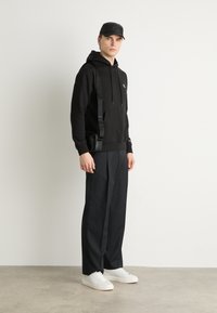 Un hoodie en coton noir avec une poche avant, associé à un pantalon large noir et des baskets blanches. Un sac bandoulière est porté sur l'épaule.