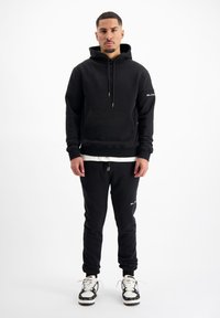 Zwarte hoodie met kangoeroezak en trekkoorden, gecombineerd met zwarte joggers met logo-accenten. Een wit shirt en patroon sneakers maken de look compleet.