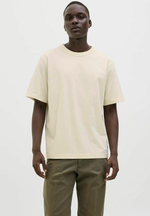 Camiseta de algodón beige con mangas cortas, escote redondo, corte holgado, diseño limpio, combinada con pantalones verde oliva, con una mínima visibilidad de marca.