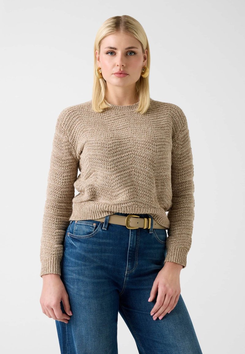 Pull beige en maille à texture ondulée, col rond et manches longues, associé à un jean en denim bleu et une ceinture beige clair.