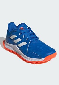 Niebieski but sportowy z teksturowaną powierzchnią, białymi paskami i pomarańczową podeszwą. Wykonany z trwałego materiału, z amortyzacją i lekką konstrukcją.