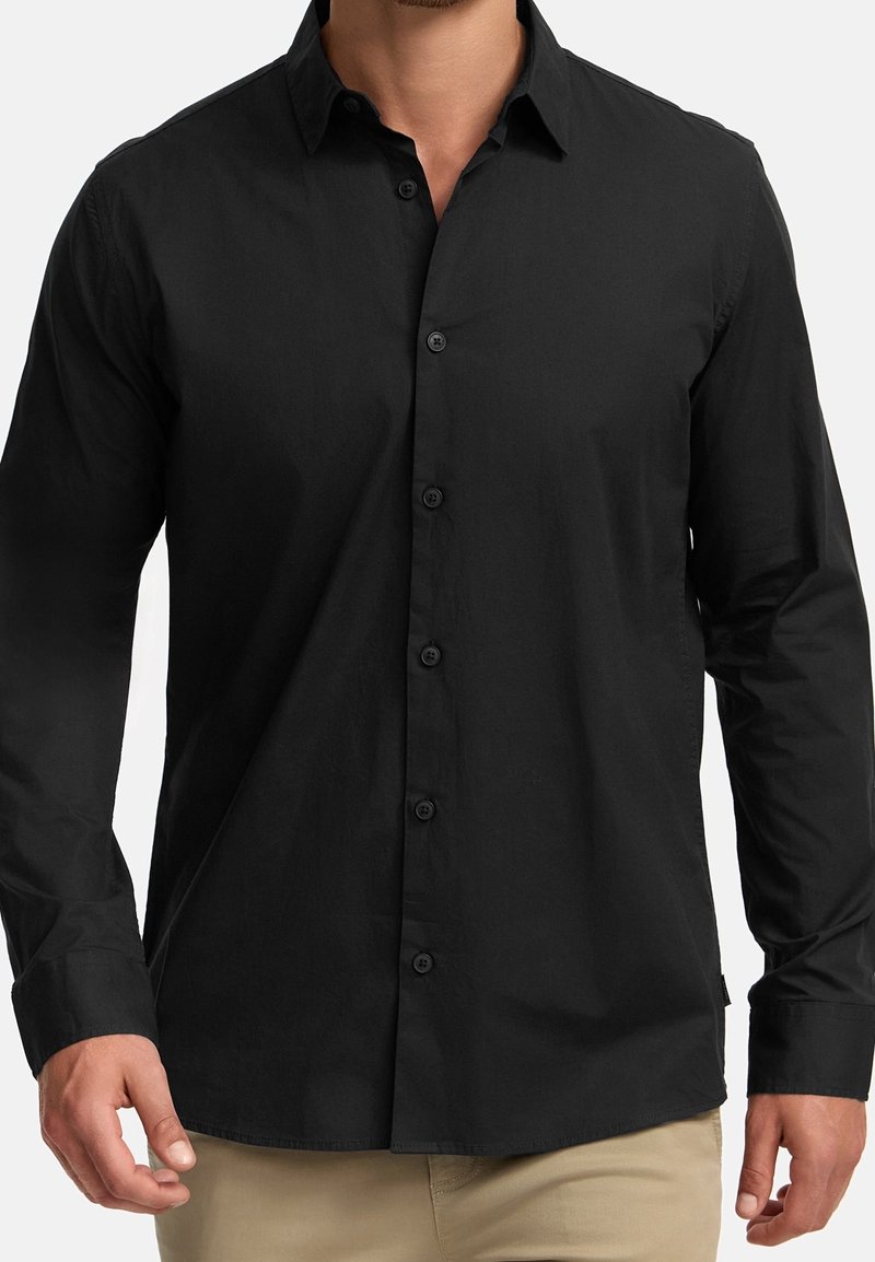 Homme portant une chemise noire ajustée à manches longues avec col et boutons, et un pantalon beige, visible du cou aux hanches.