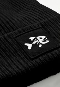 Black knit beanie with a textured pattern, featuring a woven black patch that has a white cartoon fish skeleton graphic.
