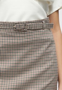 Καρό φούστα καφέ και λευκού χρώματος με ζώνη στη μέση. Το ύφασμα είναι υφασμένο και εφαρμοστό, με λεία επιφάνεια και ελαφριά γυαλάδα.