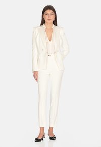 Blazer bianco su misura con revers a classe, design doppiopetto, abbinato a pantaloni slim-fit. Ballerine nere in vernice completano l'outfit.