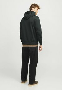 Black hoodie with a drawstring hood, worn over a tan top. Paired with loose black pants and brown shoes. Simple, minimal design.