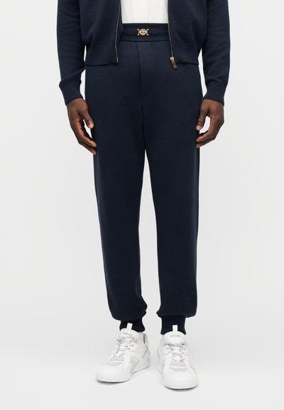 Marineblauwe joggingbroek gemaakt van zachte stof met een elastische tailleband, voorzien van een gouden logo-accent, gecombineerd met witte sneakers.