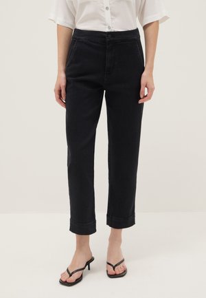 Femme portant un pantalon noir court à jambes droites avec ourlets retroussés, associé à des sandales à talons noires et une chemise blanche à manches courtes boutonnée.