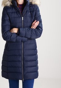 Marineblauwe pufferjas met een rits aan de voorkant, horizontale stiksels en een met bont afgewerkte kap. Beschikt over zijzakken en heeft een iets A-lijn vorm.